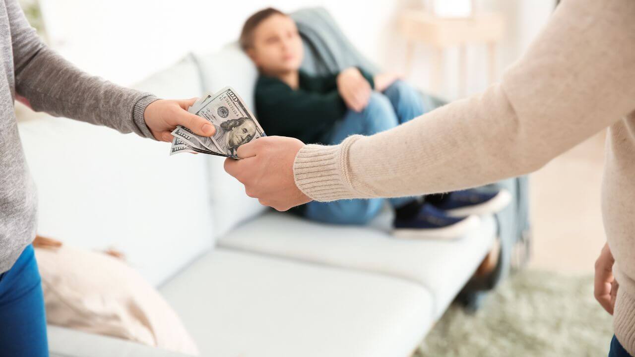 A man hands his ex-wife money while their son watches from the couch in the background.