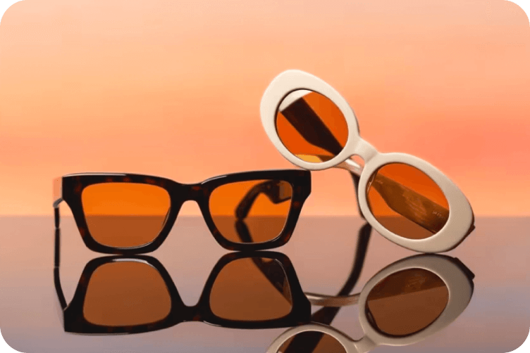 Two pairs of sunglasses with amber lenses - one black framed and one white framed - on reflective surface against orange background