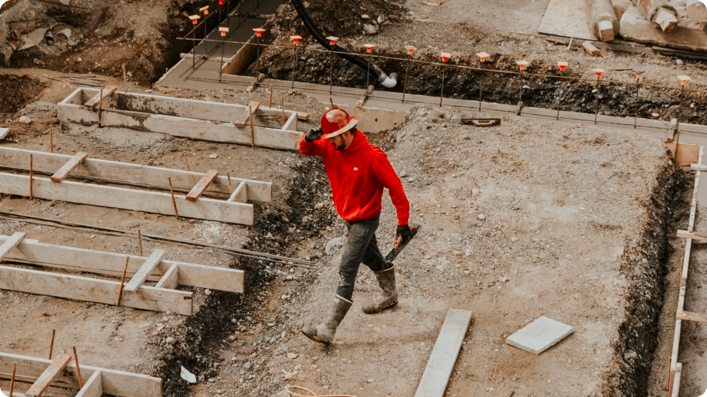 Man in a red hard hat, red hoodie, black trousers and black boots walking across a construction site