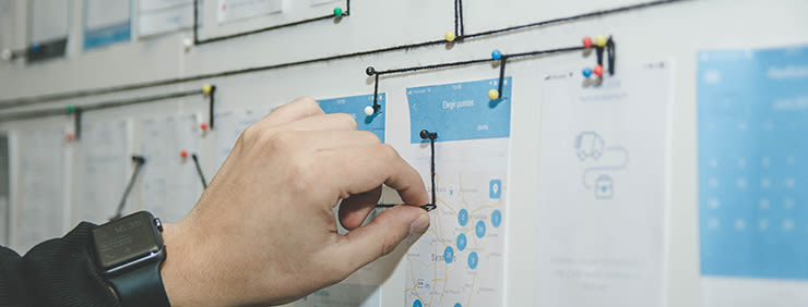 Hand connecting points on a map diagram with a black string, pinned to a whiteboard with pushpins, with a smartwatch visible on the wrist.