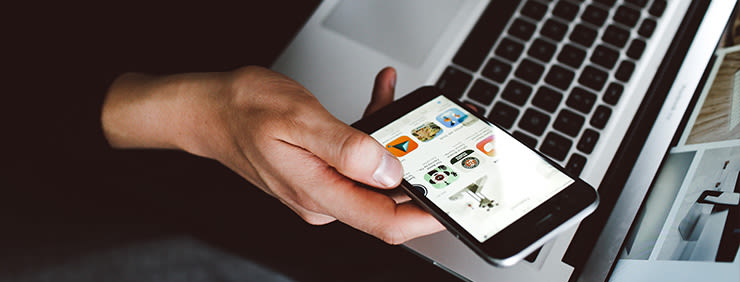 A hand holding a phone with the App Store open, resting on a laptop.