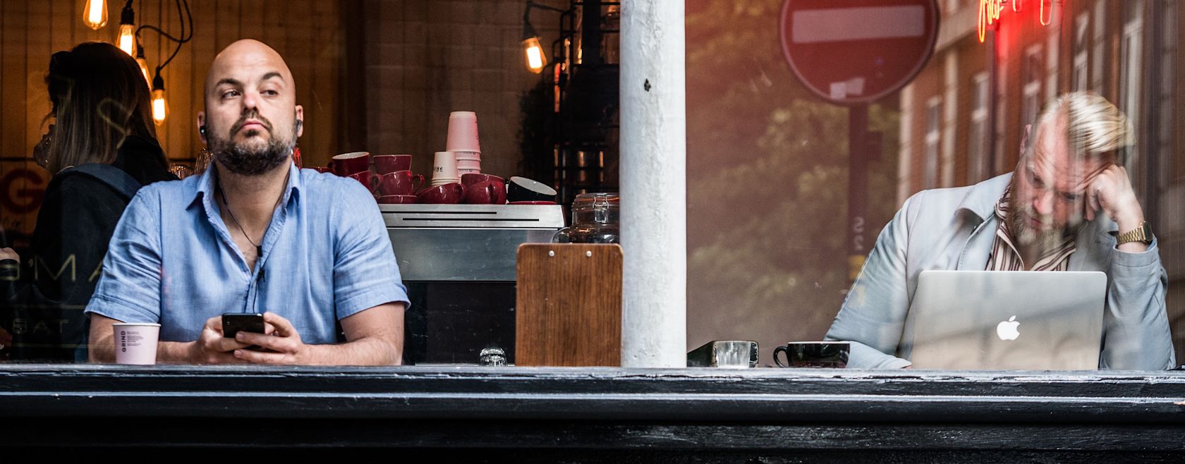 Two men seen through a cafe window; one is bald, holding a phone, and the other is using a laptop.