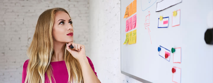 A woman with long blonde hair, wearing a pink dress, contemplates a whiteboard covered in colorful sticky notes and drawn diagrams. She's holding a marker in her hand.