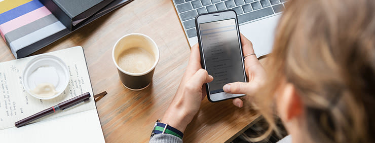 Person using a smartphone while sitting at a desk with a laptop, notebook, pen, coffee, and small container.