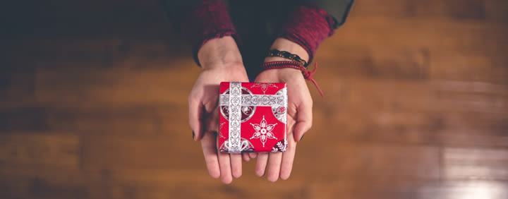 A gift wrapped in red paper with white snowflake patterns held in cupped hands against a wooden floor.