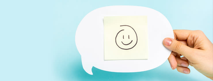 Hand holding a white speech bubble with a yellow sticky note featuring a simple smiley face drawing.