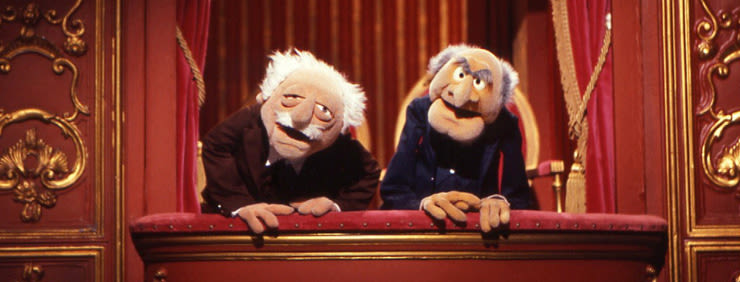 Statler and Waldorf, two elderly Muppet characters, sitting in a theater balcony with red drapes and gold ornamentation, ready to heckle the show.