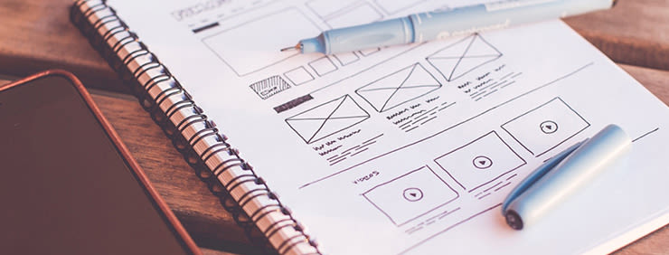 Notebook with a hand-drawn website wireframe and pen on a wooden table, with a smartphone visible on the side.