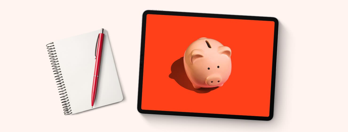 Notebook and pen next to a tablet displaying a piggy bank on an orange background.