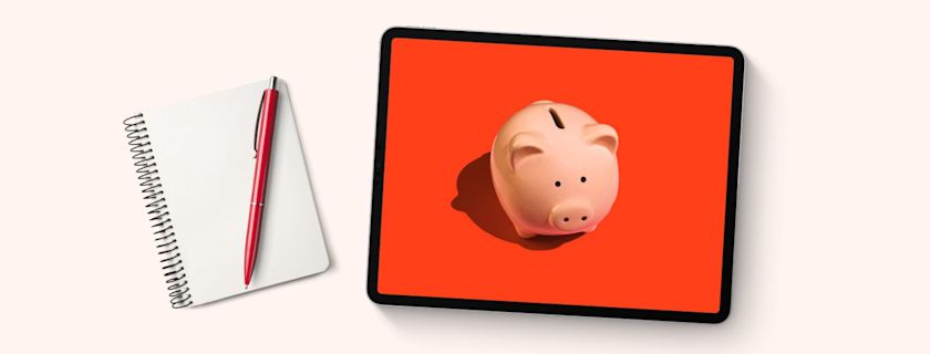 Notebook and pen next to a tablet displaying a piggy bank on an orange background.