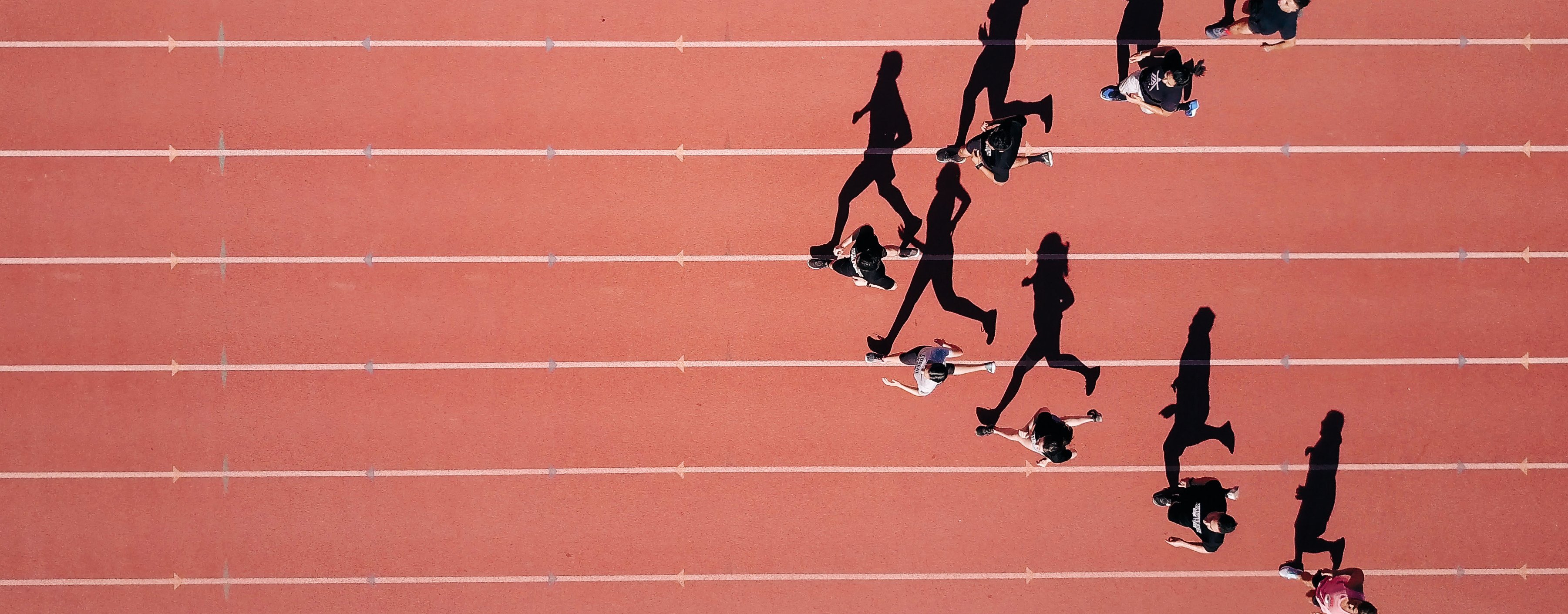 Aerial view of people running on a red track with white lines, casting long shadows.