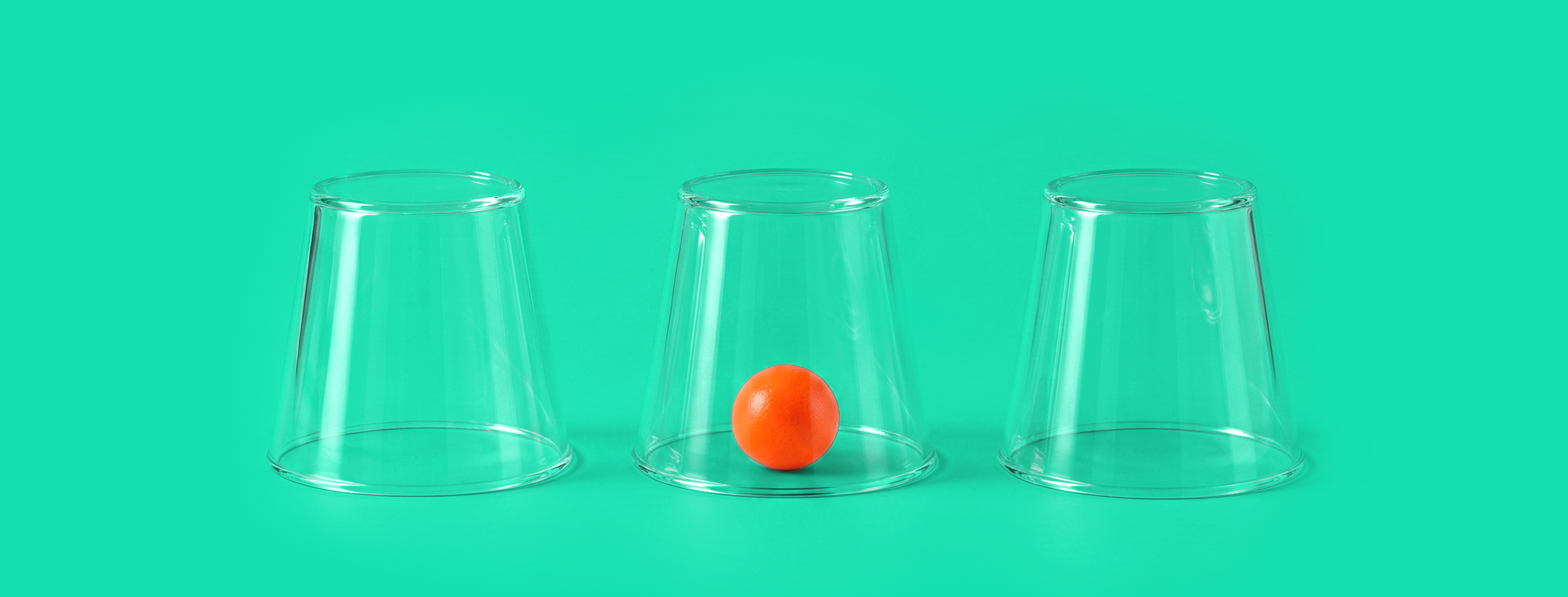 Three transparent cups upside down on a green background, with a small orange ball visible under the middle cup.