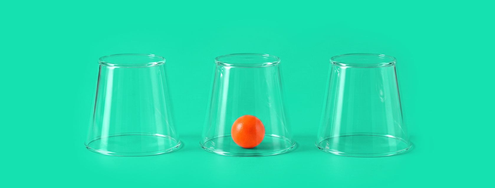 Three transparent cups upside down on a green background, with a small orange ball visible under the middle cup.