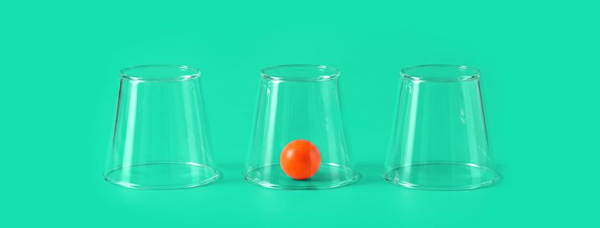 Three transparent cups upside down on a green background, with a small orange ball visible under the middle cup.