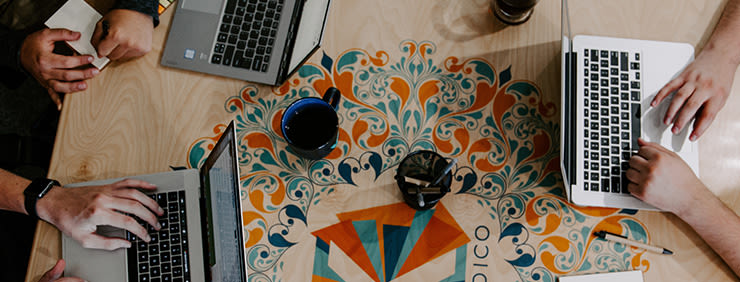 Overhead view of four hands working on three laptop computers at a patterned table. A coffee mug, pen holder and notebook are also on the table.