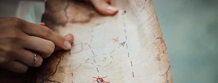 A hand with a ring points to a spot on an old-looking map with an 'X' marking the location.
