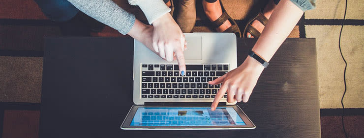 Overhead shot of two people pointing at a laptop and tablet on a dark wooden table.