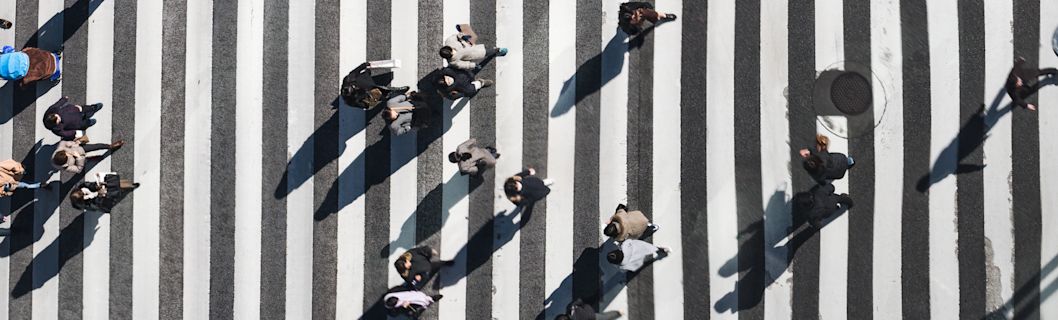 Overhead view of a pedestrian crosswalk with many people crossing it. The crosswalk is black and white, and the people are casting shadows.