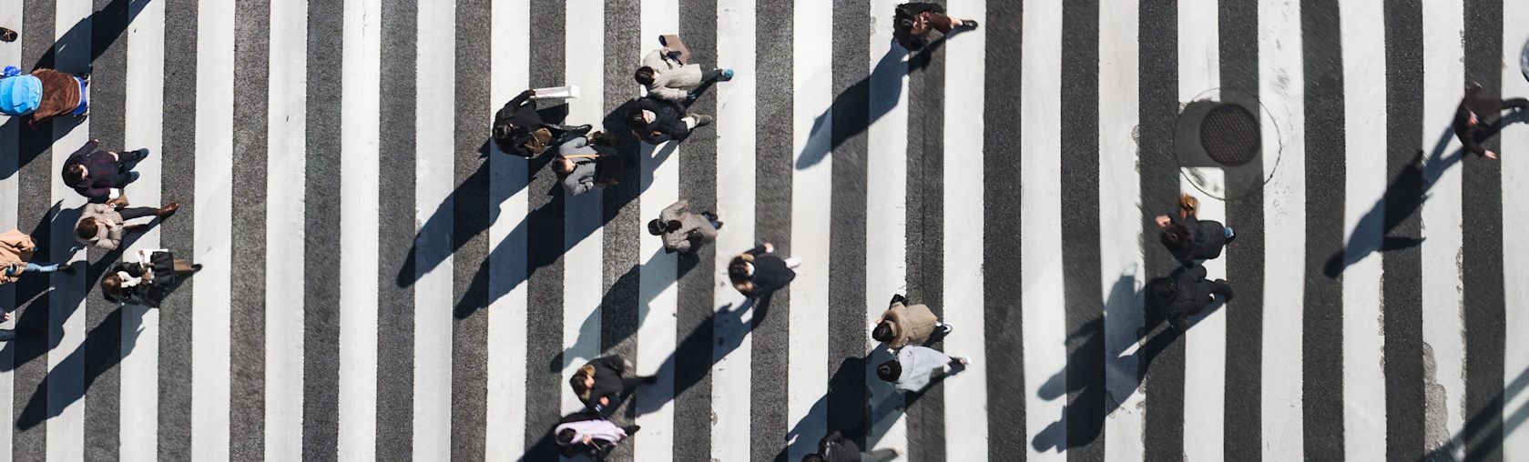 Overhead view of a pedestrian crosswalk with many people crossing it. The crosswalk is black and white, and the people are casting shadows.