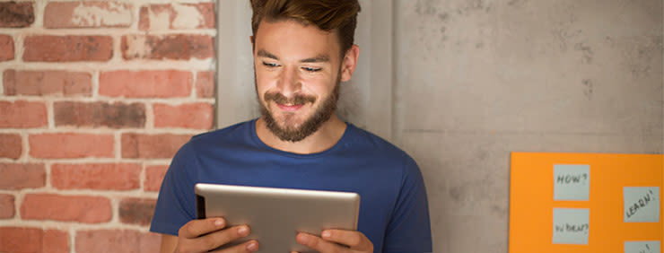Man with a beard looking at a tablet in front of a brick wall and a bulletin board.