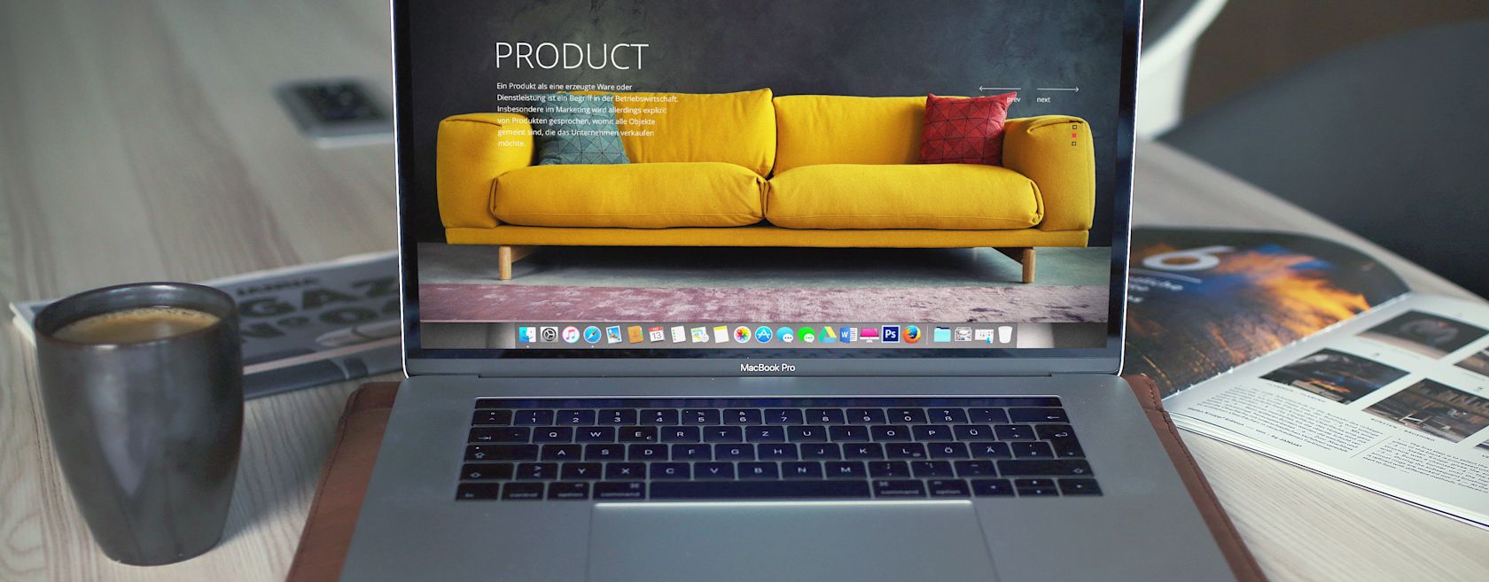 A MacBook Pro displays a product landing page with a yellow couch and a red pillow, resting on a wooden surface with a coffee mug and open magazine nearby.