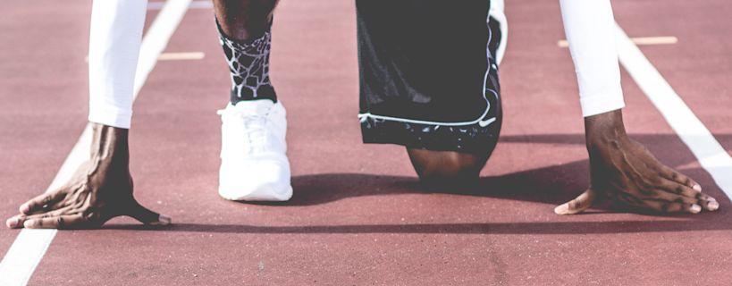 A person in a starting position on a running track, hands on the track, with a white shoe and black shorts visible.