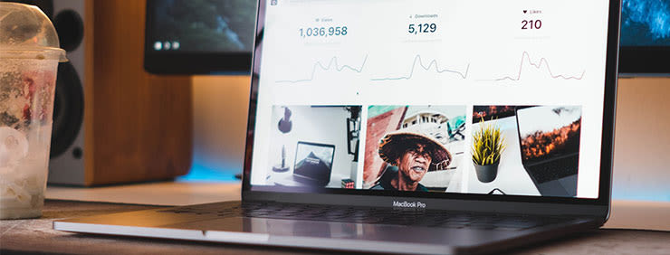 A MacBook Pro displaying website analytics with views, downloads, and likes, with a drink and other tech in the background.