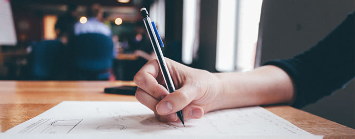 Hand holding a pen and writing/drawing on paper at a desk in a cafe.