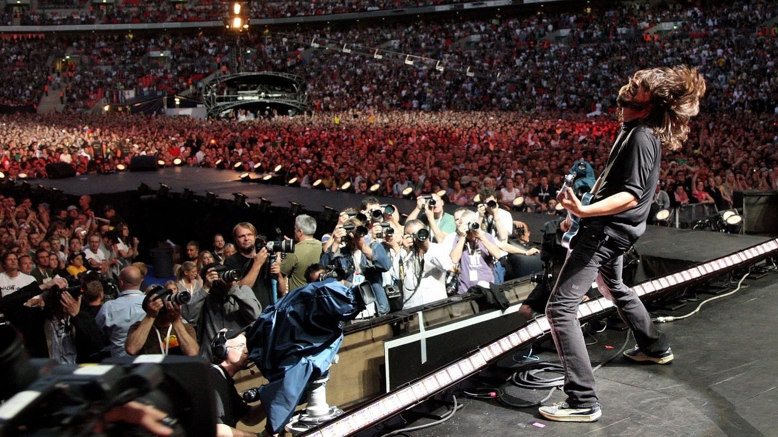 A musician plays guitar on stage in front of a large crowd, with many photographers taking pictures.