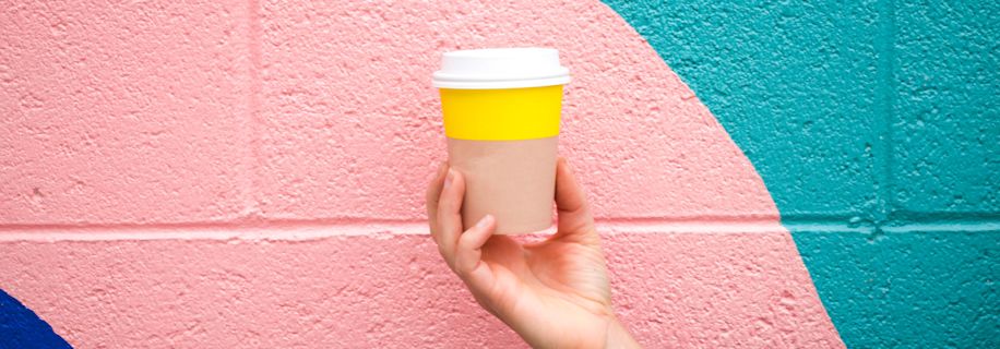 A hand holding a colorful coffee cup in front of a painted brick wall. The cup has a white lid, yellow middle and tan bottom. The wall is painted in pink, teal, and blue.