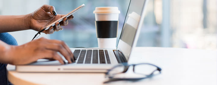 Person using a laptop while holding a smartphone, with a coffee cup and glasses on the table.