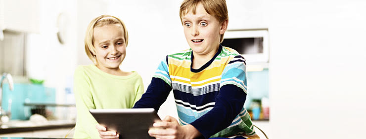 Two young children looking at a tablet, one smiling, one with a surprised expression.