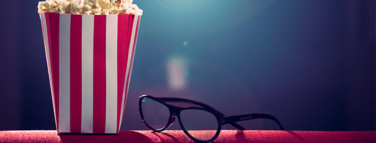 A striped bucket of popcorn and 3D glasses on a red surface, suggesting a movie viewing experience.