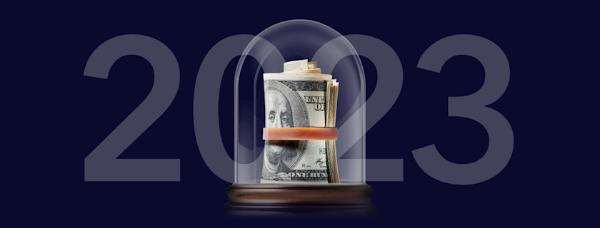"2023" text overlaying a roll of US currency, secured with a band and displayed under a glass dome. The background is a dark blue.