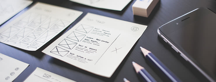 Sketches of website layouts alongside pencils and a phone on a dark surface, suggesting design and planning.