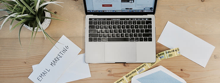 Laptop, envelopes, labels, and a plant on a wooden desk, with "EMAIL MARKETING" handwritten on one envelope.