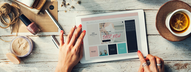 Woman shopping online on a tablet with makeup and tea nearby.