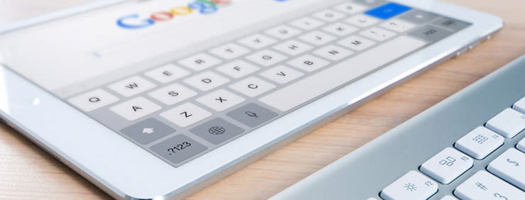 A tablet displaying a Google search bar and keyboard, with a physical keyboard partially visible next to it.