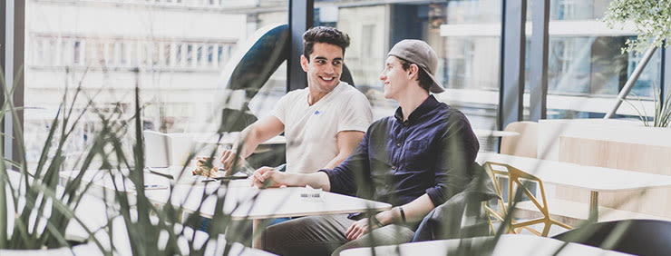 Two friends are sitting at a table, talking and smiling, in a modern cafe. One wears a white t-shirt, the other a button-down shirt and baseball cap.
