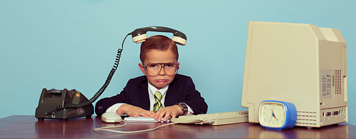 Frustrated young boy dressed as a businessman with a phone receiver on his head, sitting at an old-fashioned office desk with vintage computer and rotary phone.
