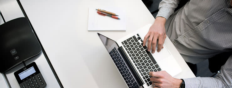 Overhead shot of a person working on a laptop at a white desk with a Cisco phone, notepad, and pens.