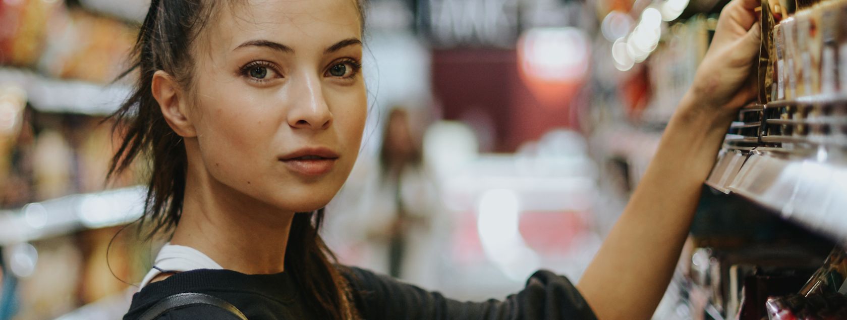 A young woman with green eyes and brown hair pulled back, looking at the camera while reaching for something on a shelf in a store.