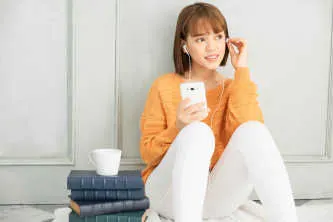Teenage girl with headphones using mobile phone while sitting against a wall