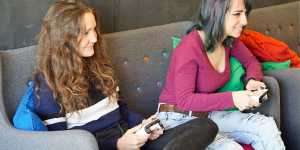 two young women sitting on a sofa playing video games and having fun while starring at a screen and holding the console controllers in their hands