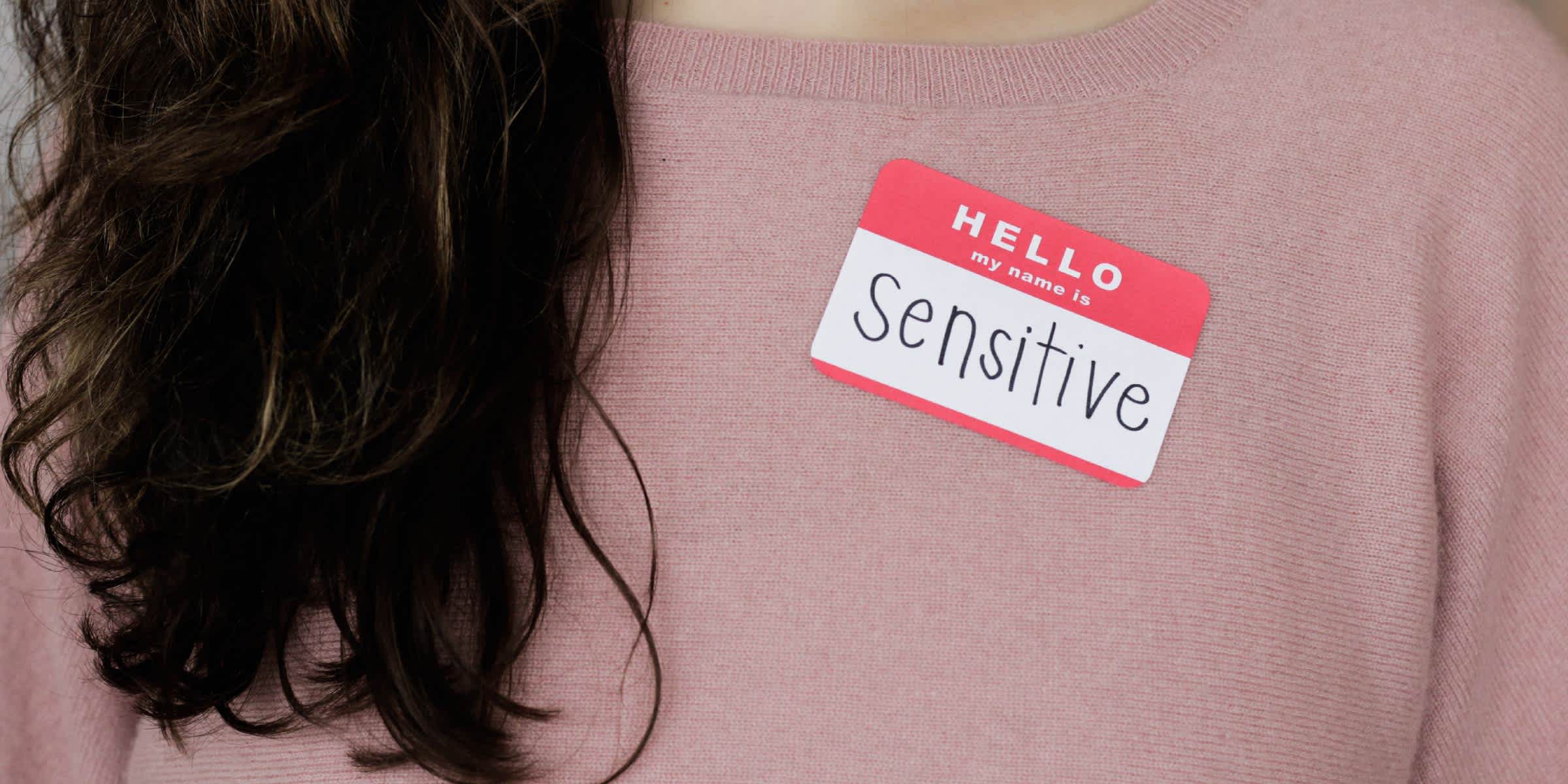 close up photo of a pink sweater worn by a person covered by brown hair and with a sticker attached to it stating hello my name is sensitive