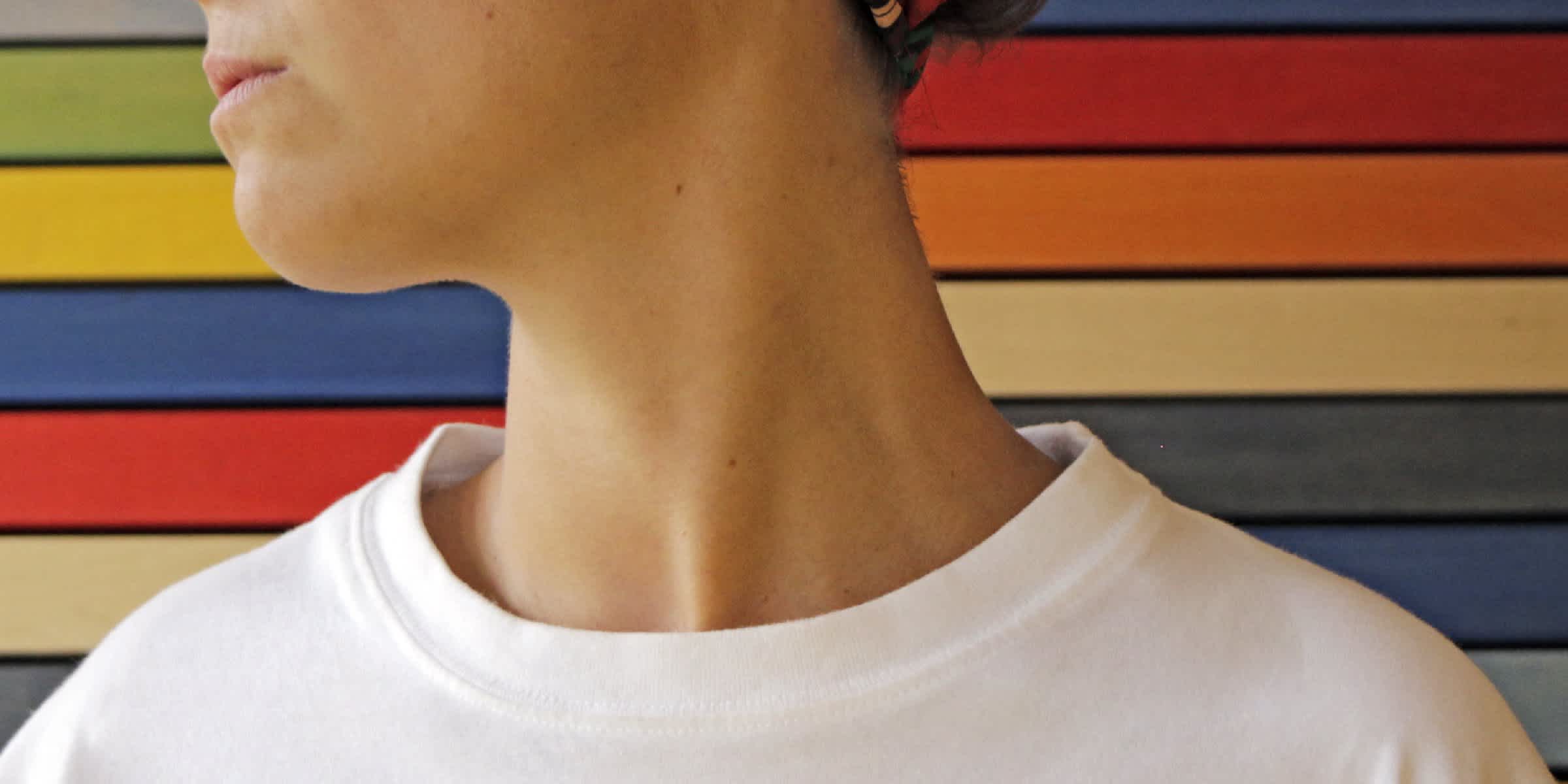 A closeup photograph of a person's neck, against a rainbow background, wearing a white t-shirt