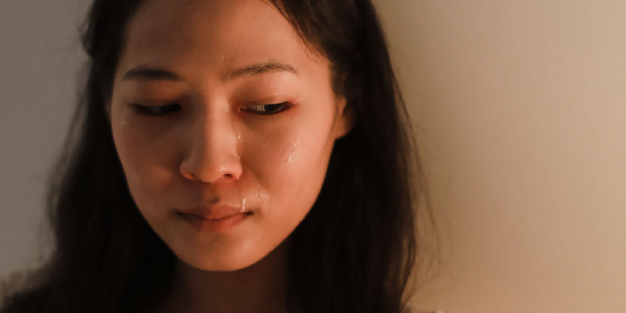 Close-up of a young woman with long dark hair looking down, tears streaming down her cheek and catching the warm, dim light. Her lips are pressed together, and her brow is slightly furrowed, conveying quiet distress, against a soft beige wall.
