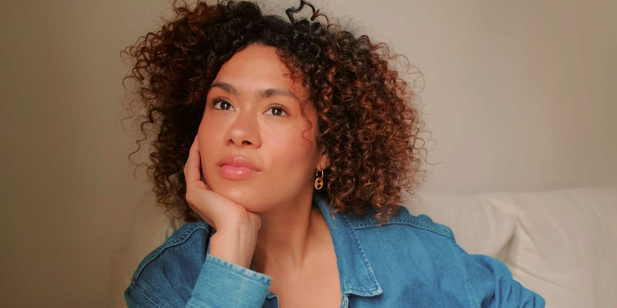 A woman in a denim shirt sits on a sofa with her chin resting in her hand, looking into the distance with a pensive expression.