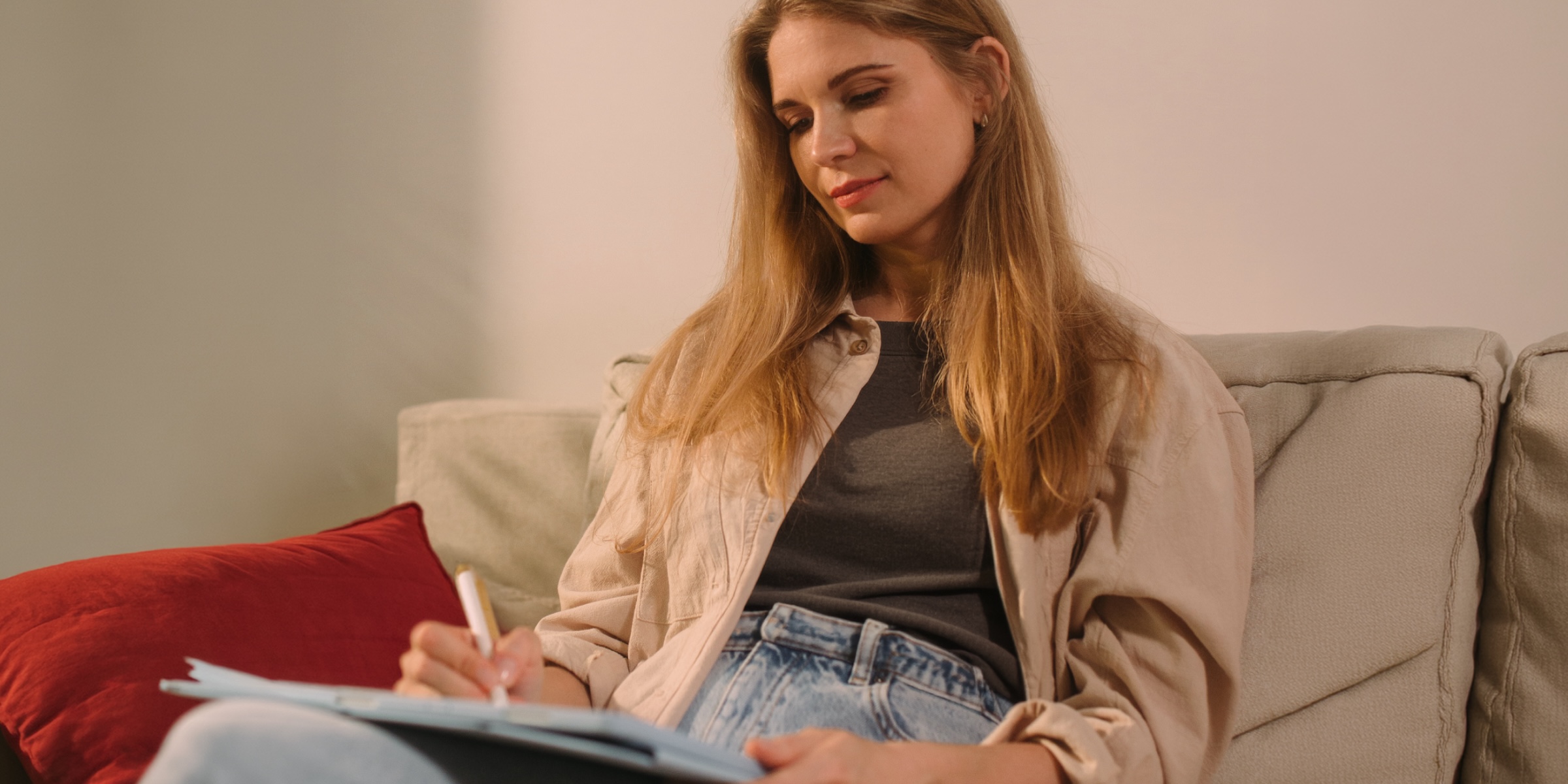 A woman with long hair sits relaxed on a beige sofa beside a red cushion, writing notes on a clipboard as she prepares for an appointment.