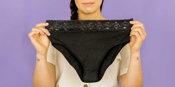 A woman holds up a pair of Flux black bikini-style period underpants, with lace trim on the waistband. 